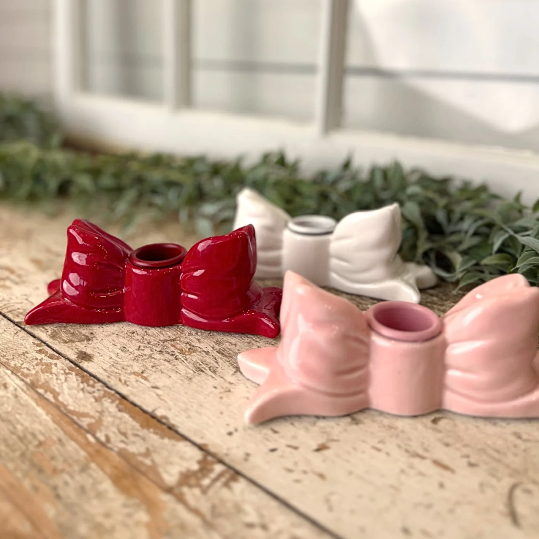 Two stoneware bow-shaped candle holders, one red and one pink, on a wooden surface with greenery in the background.
