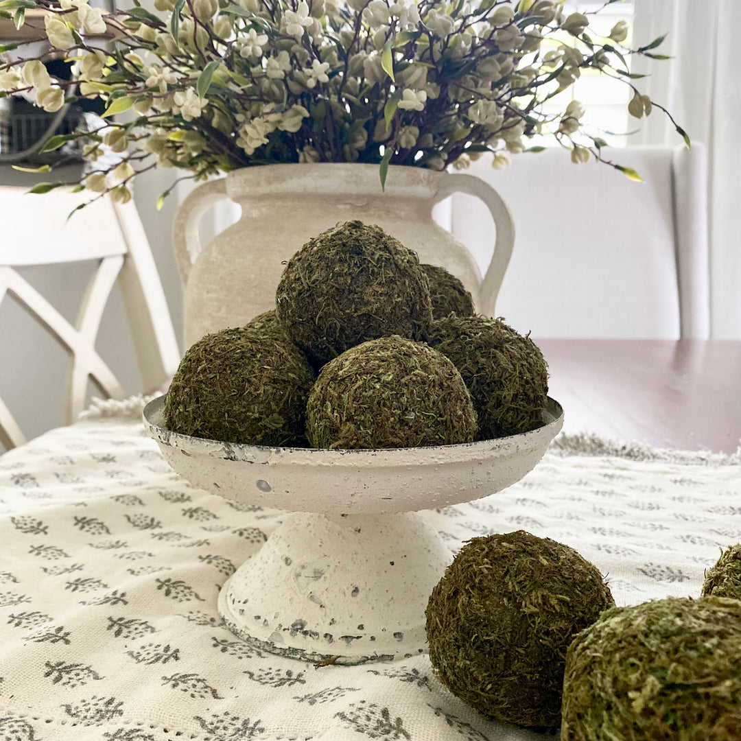 A distressed white metal pedestal plate with a textured, vintage-inspired finish, displayed on a table with decorative moss balls on top.