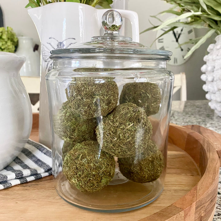 A glass jar on a wooden surface containing spherical dried moss balls used as bowl fillers.