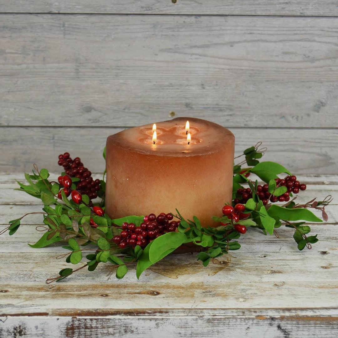 Boxwood & Berries Candle Ring with a brown wax candle surrounded by green artificial foliage and red artificial berries, placed on a white wooden surface.