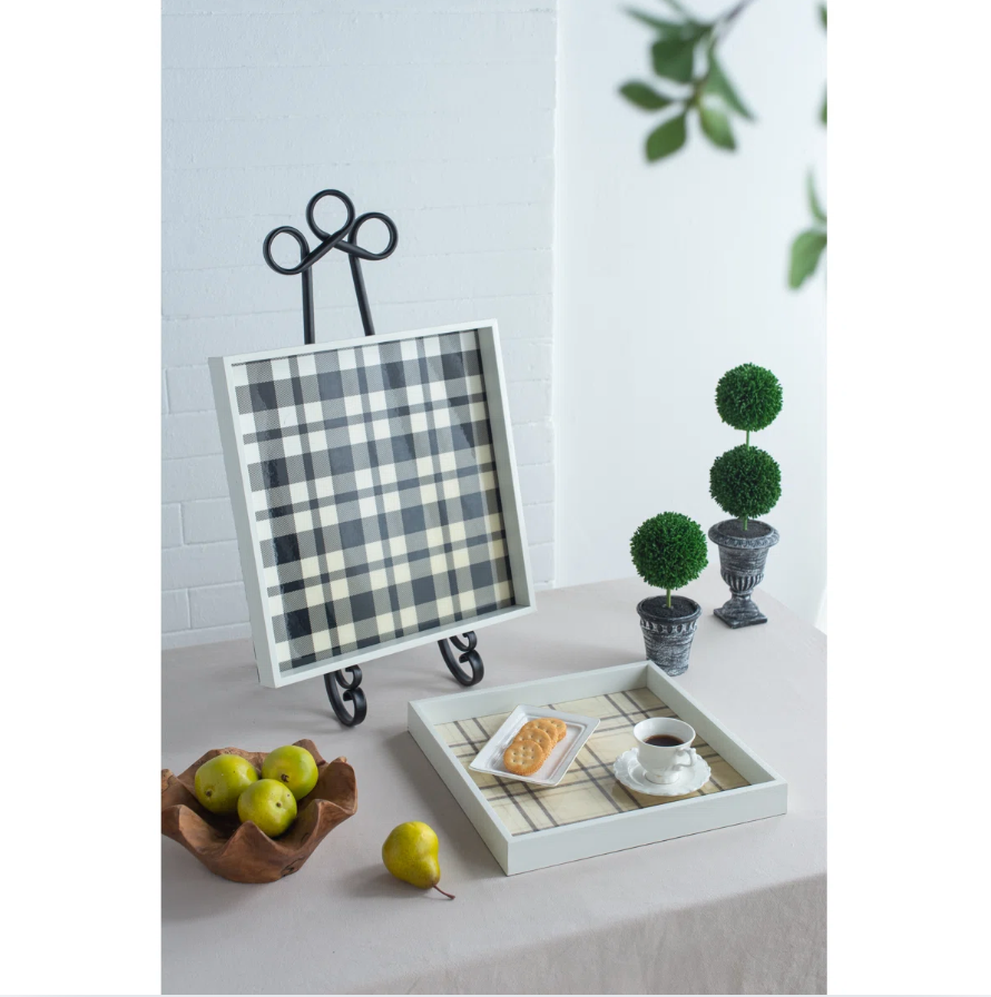 Buffalo check and plaid decorative trays displayed on a small easel stand on a table, styled with a tray, small potted topiary trees, a bowl of green apples, and a cup of coffee in a bright indoor setting.