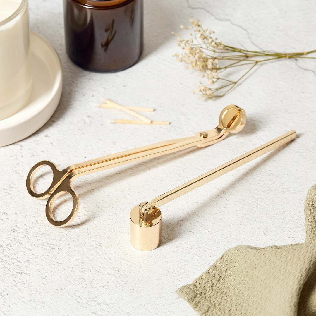 Gold candle wick trimmer and candle snuffer on a textured surface with a candle and flowers in the background