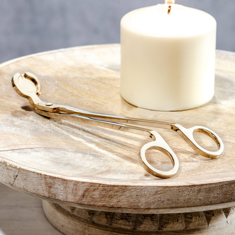 A gold-colored metal candle wick trimmer resting on a wooden surface next to a lit candle.
