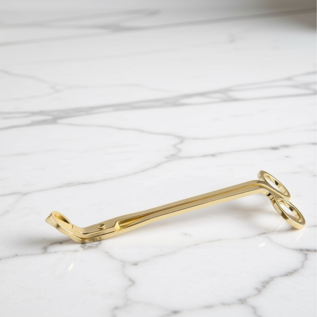 A polished gold metal candle wick trimmer resting on a white marble surface with gray veining.