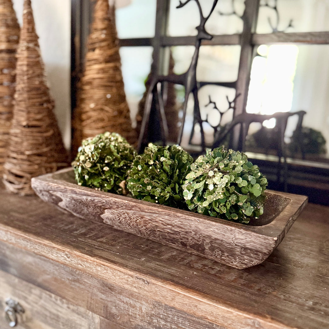 Decorative arrangement with green spherical bowl filler in a carved wooden tray on a wooden surface.