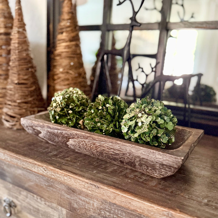 Decorative arrangement with green spherical bowl filler in a carved wooden tray on a wooden surface.