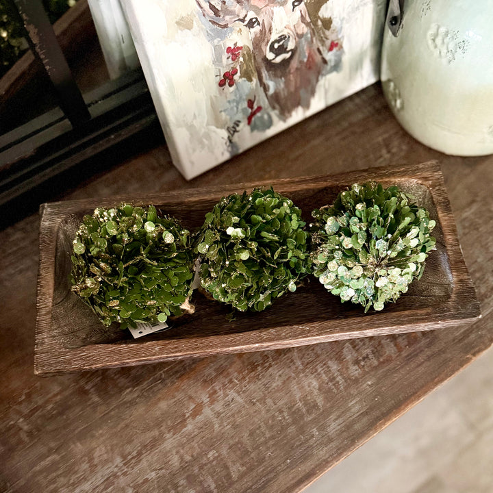 Three green decorative ball ornaments in a carved wooden tray on a wooden surface with a painting in the background.