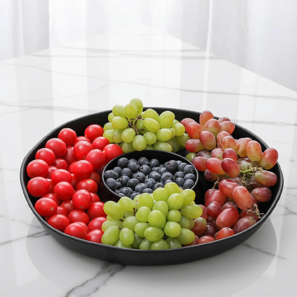 A round black chip and dip serving platter filled with assorted fruit, including red cherry tomatoes, green and red grapes, and blueberries, set on a white marble countertop.