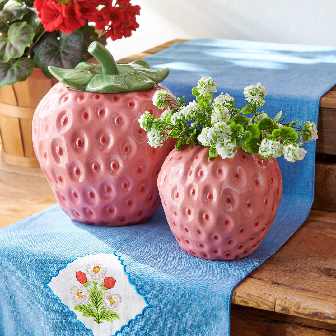 Two pink strawberry-shaped planters with flowers on a blue tablecloth.