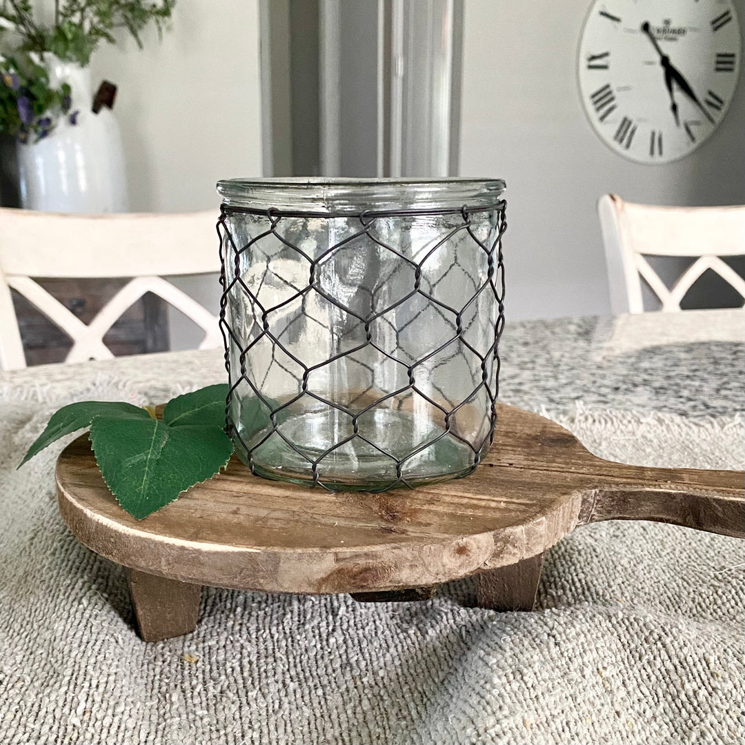 A clear glass candle holder with chicken wire, placed on a wooden board, resting on a table with a clock in the background.
