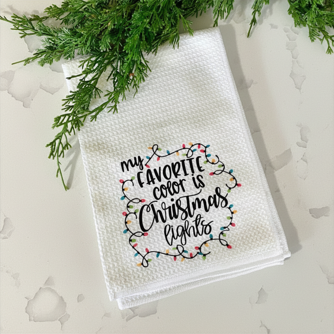 White textured towel with 'my favorite color is Christmas lights' text and Christmas lights design, placed on a light background with greenery.