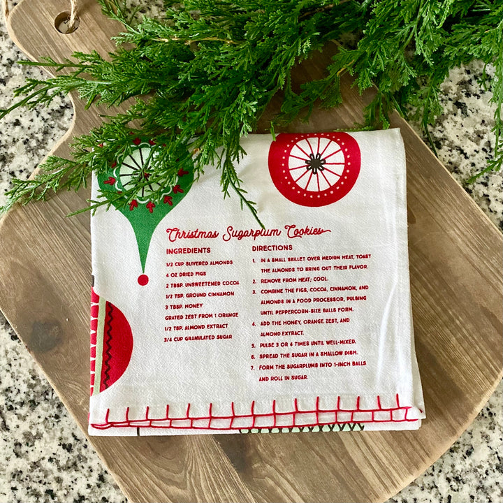 A white cotton kitchen towel with red text and decorations, featuring a Christmas sugarplum cookie recipe, placed on a wooden board with green pine branches in the background.