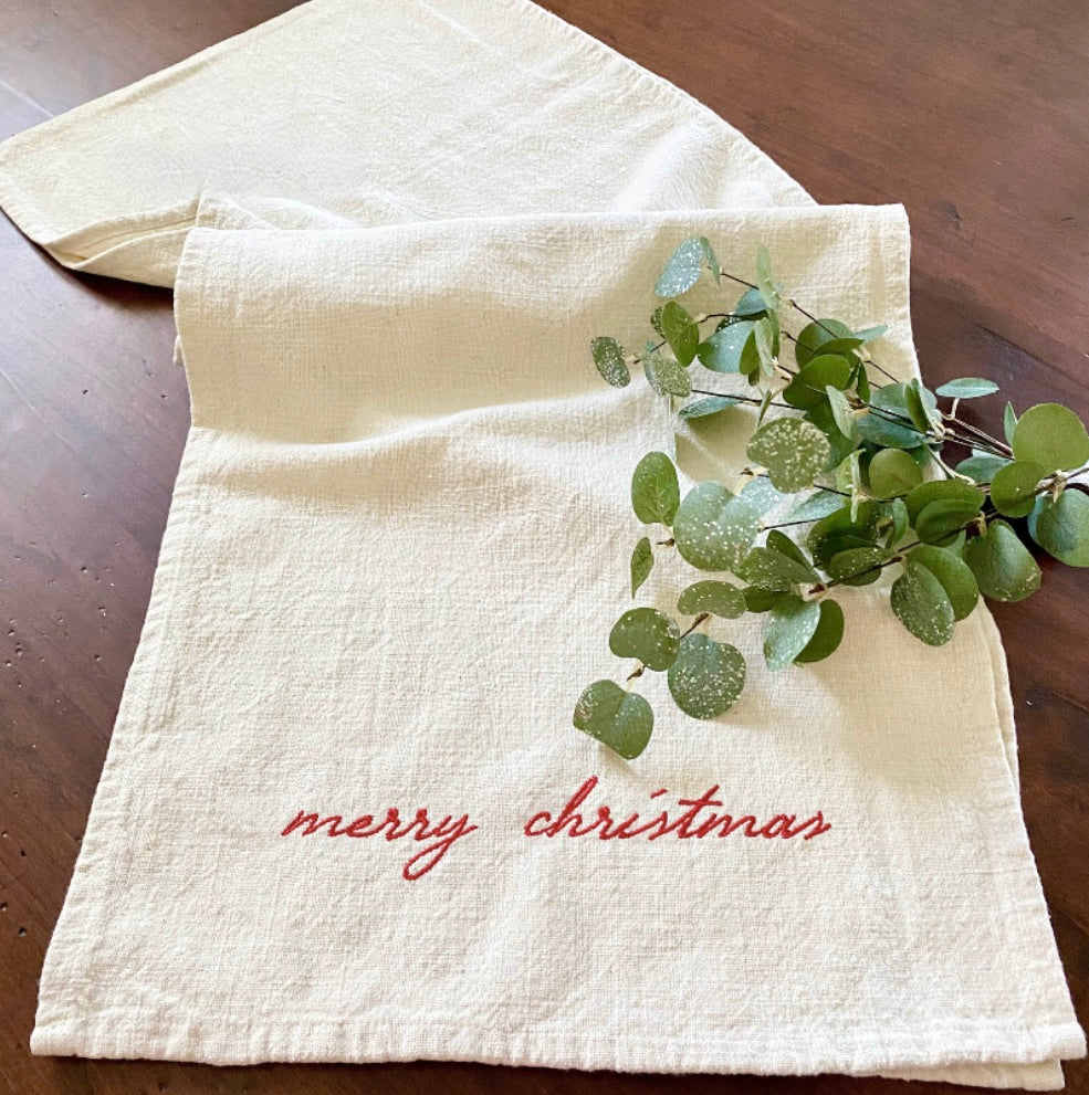 A cream-colored Christmas table runner with 'Merry Christmas' embroidered in red, accompanied by a small greenery decoration, placed on a wooden surface.
