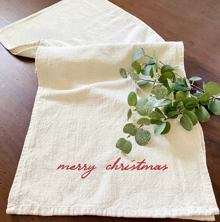 A cream-colored Christmas table runner with 'Merry Christmas' embroidered in red, accompanied by a small greenery decoration, placed on a wooden surface.