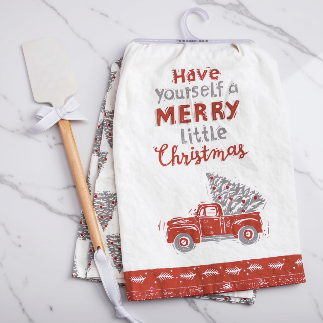 A white kitchen tea towel with a Christmas print hangs on a hanger on a white marble surface, next to a wooden-handled spatula. The towel features the text 'Have yourself a MERRY little Christmas' above a red vintage truck carrying a Christmas tree, with a red patterned border at the bottom.