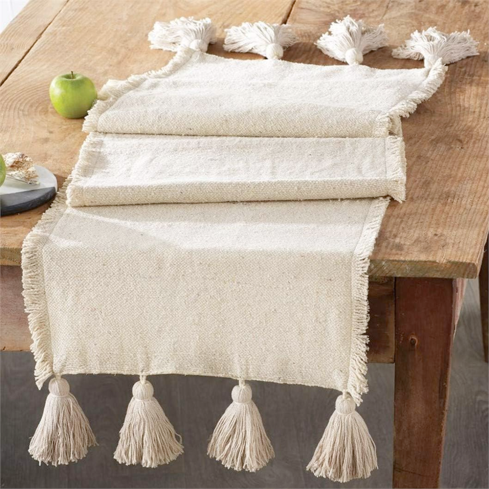 An ivory cotton table runner with fringe trim and large tassels is displayed on a wooden table. The runner features a subtle woven sparkle pattern.