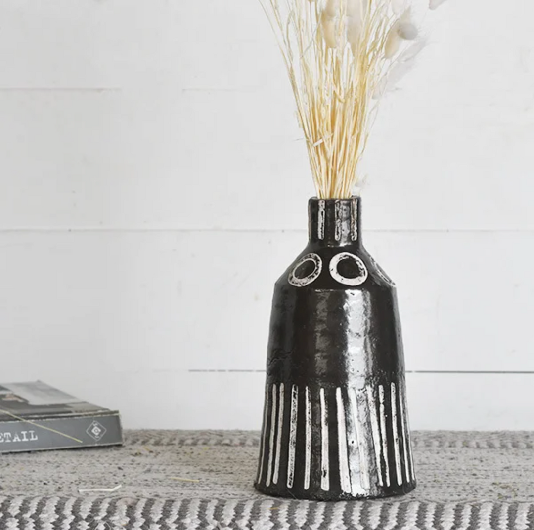 A black and white striped vase with a geometric pattern, displaying a bundle of dried grasses, placed on a flat surface with a book in the background.