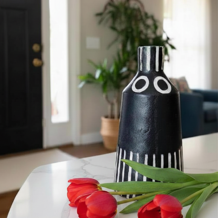 Black and white vase with red tulips on a table in a room with plants and furniture.