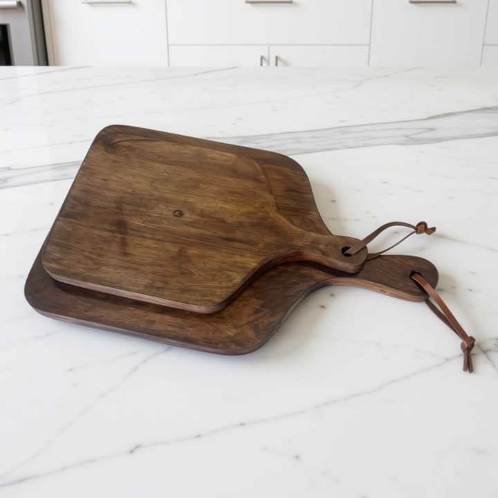 Set of two walnut brown wooden serving boards with leather handles resting on a white marble kitchen island.