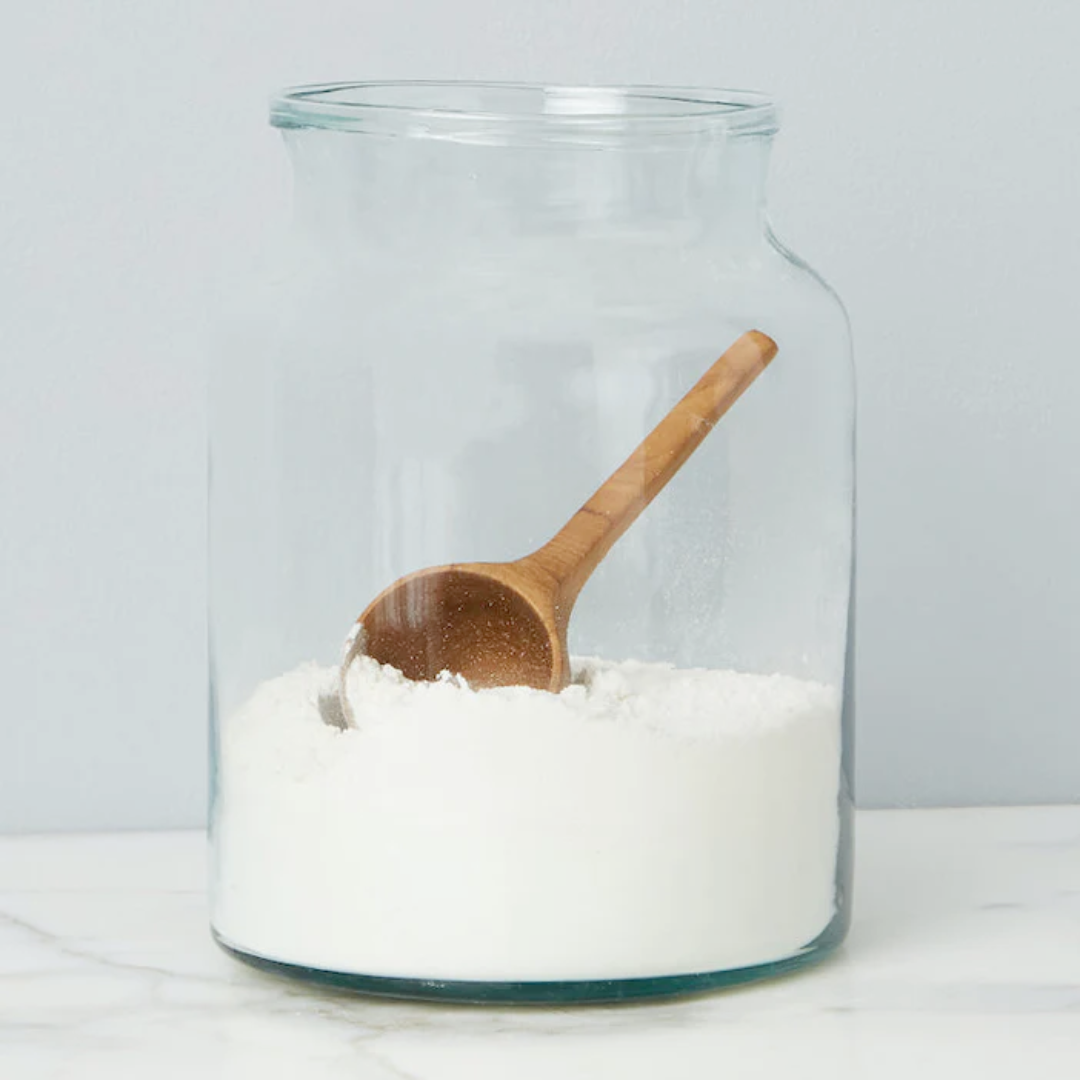 A clear glass mason jar with a wooden spoon inside, containing white granular substance, placed on a white surface.