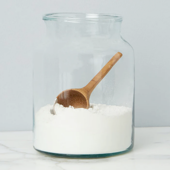 A clear glass mason jar with a wooden spoon inside, containing white granular substance, placed on a white surface.