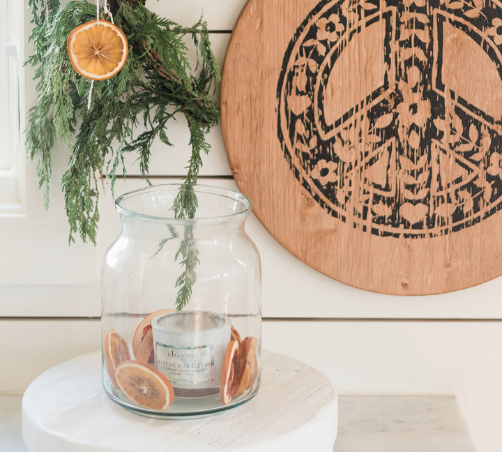 A clear mason jar with a candle inside and dried orange slices around the base, placed on a white surface with a decorative wooden board in the background.