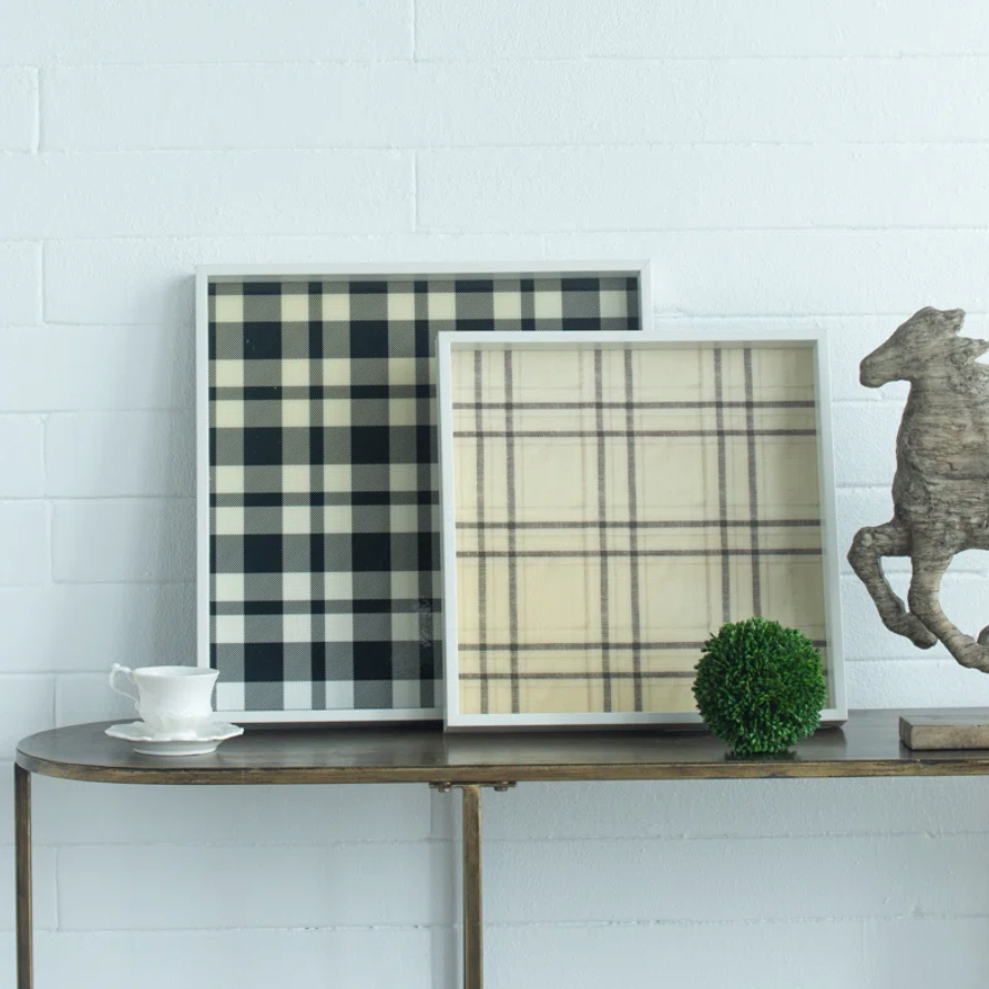 Set of two plaid decorative trays leaning on a console table against a white brick wall. One frame contains a black and cream buffalo plaid pattern, and the other contains a beige and brown plaid pattern.