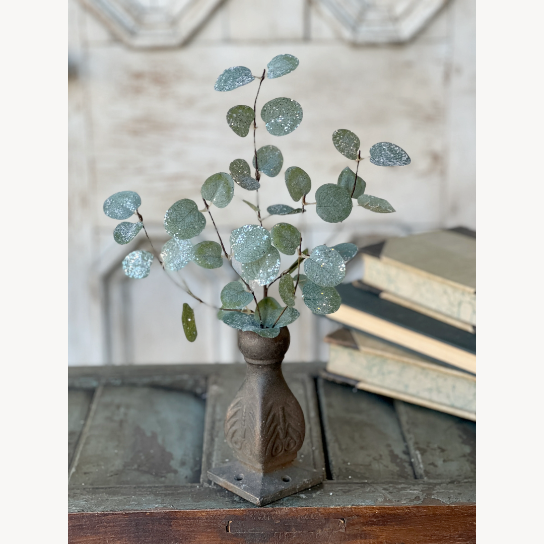 A decorative eucalyptus pick with rounded leaves dusted with glitter, resembling freshly fallen snow, placed in a vase with a rustic background.