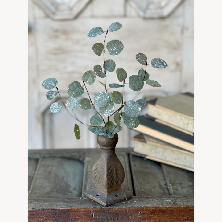 A decorative eucalyptus pick with rounded leaves dusted with glitter, resembling freshly fallen snow, placed in a vase with a rustic background.