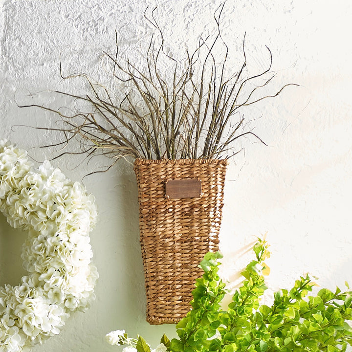 A wicker vase filled with artificial curly willow branches, positioned against a white wall with a small inset of green foliage.