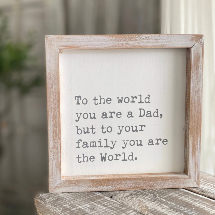 A white block sign with weathered wood frame containing the text 'To the world you are a Dad, but to your family you are the World.'.