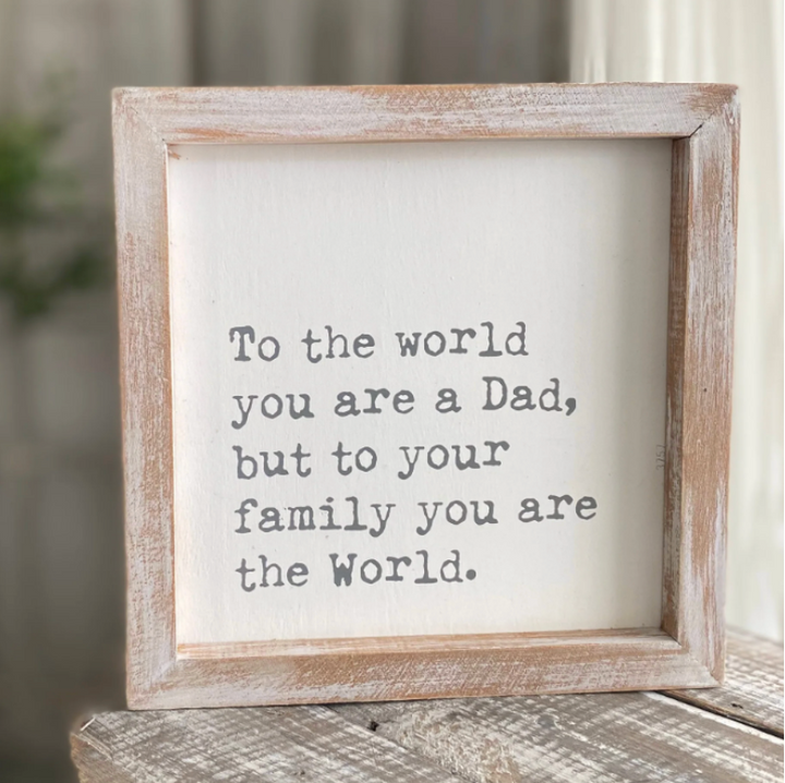 A white block sign with weathered wood frame containing the text 'To the world you are a Dad, but to your family you are the World.'.