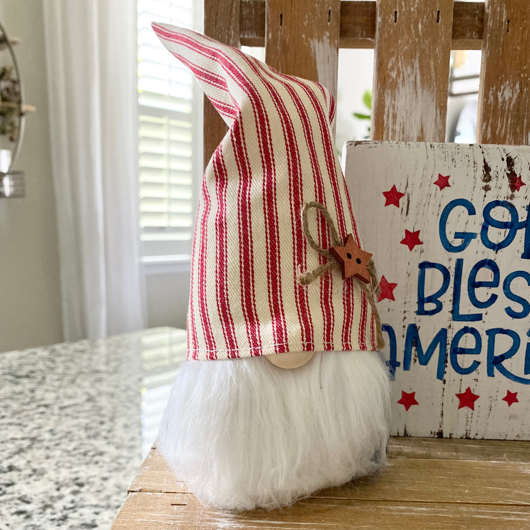 A decorative Americana themed arrangement featuring a wooden star, a gnome with a blue star hat, a small American flag, and a 'God Bless America' sign, all placed on a tiered tray.