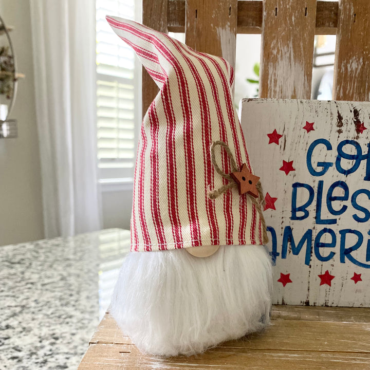A decorative Americana themed arrangement featuring a wooden star, a gnome with a blue star hat, a small American flag, and a 'God Bless America' sign, all placed on a tiered tray.