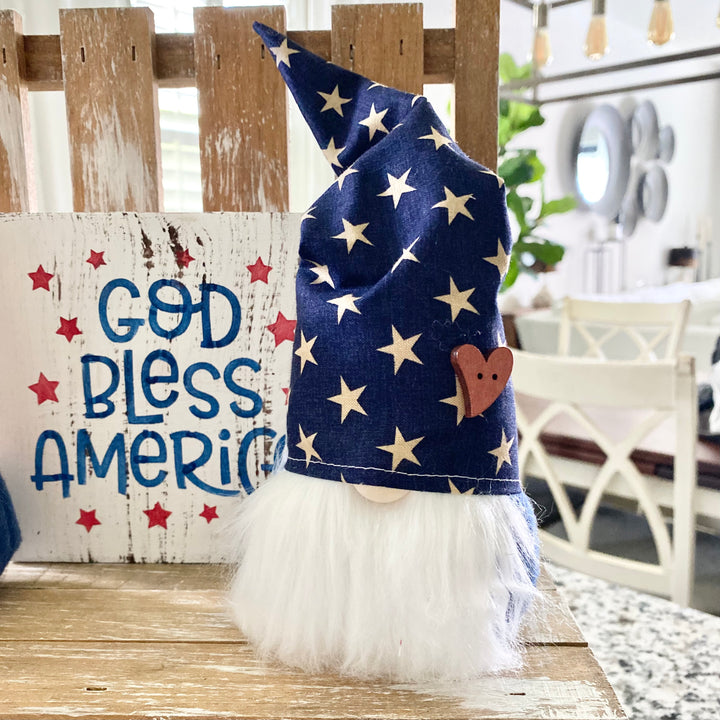 A decorative Americana themed arrangement featuring a wooden star, a gnome with a blue star hat, a small American flag, and a 'God Bless America' sign, all placed on a tiered tray.
