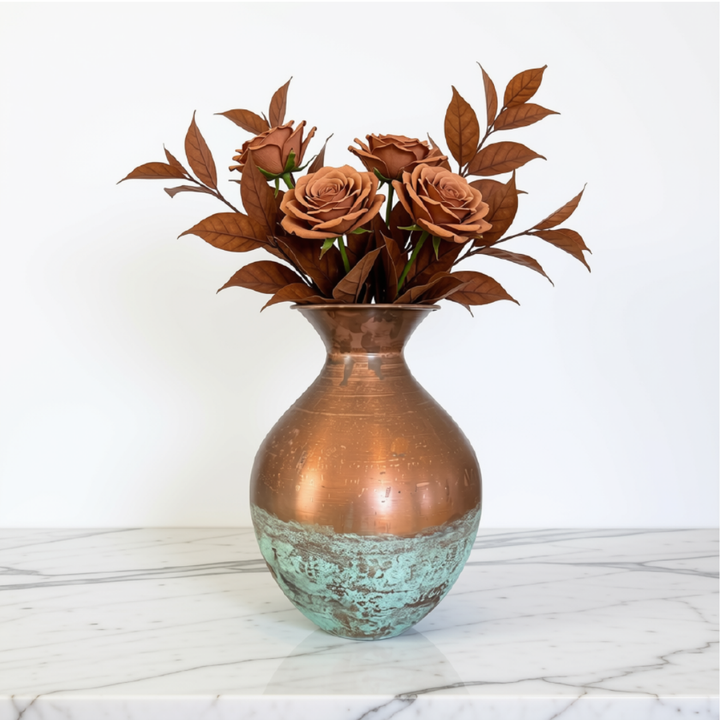 A decorative metal vase with a distressed copper upper body and a turquoise-green lower section, filled with artificial brown roses and leaves, set on a white marble surface against a white wall.