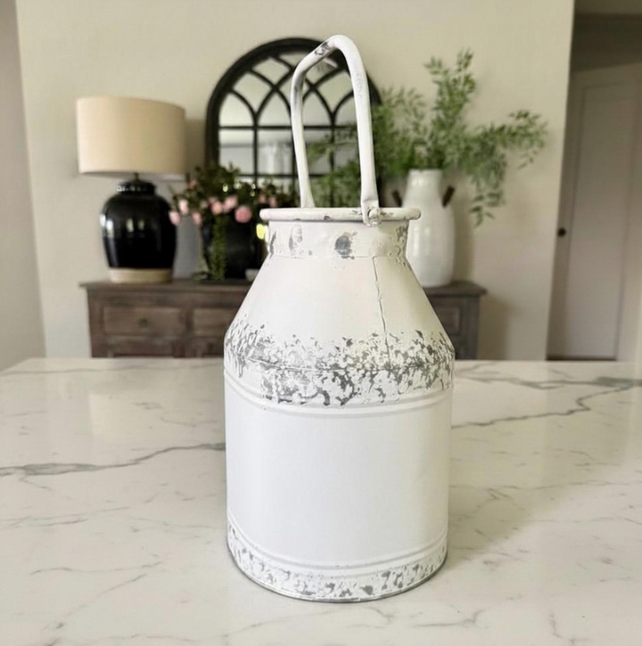 A distressed white metal milk can decorative piece with a handle, set on a white marble countertop in a bright, modern room.