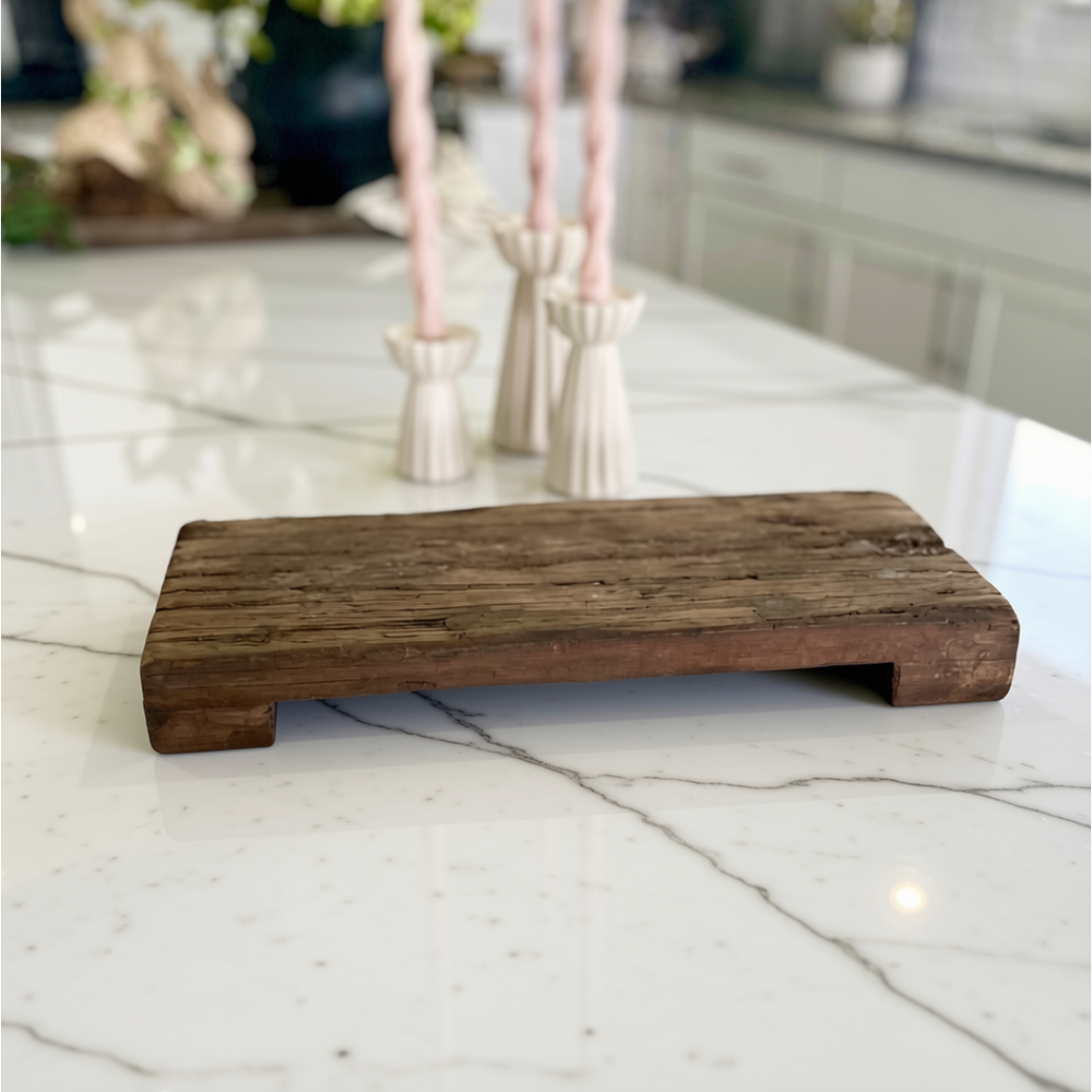 A rustic rectangular wooden serving board or tray with a low raised edge, shown on a white marble countertop in a kitchen setting with blurred pink candles in the background.