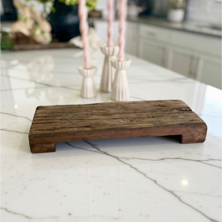 A rustic rectangular wooden serving board or tray with a low raised edge, shown on a white marble countertop in a kitchen setting with blurred pink candles in the background.