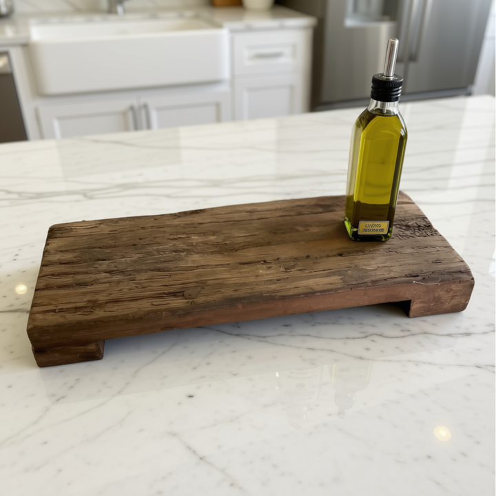 A rustic rectangular wooden serving board with a small glass oil bottle placed on top, set on a white marble kitchen countertop with a blurred kitchen background.