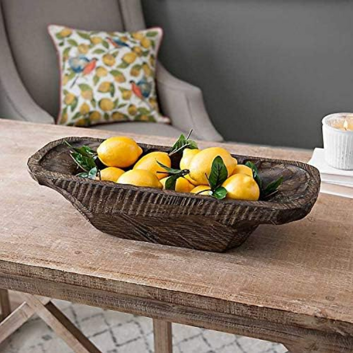 A Hand-Carved Wooden Dough Bowl features a rich dark brown finish with distressed detailing and visible hand-tool marks filled with yellow lemons and green leaves, placed on a rustic wooden table in a living room setting.