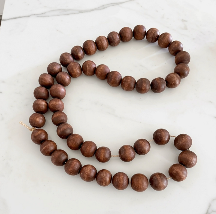 A strand of medium brown wooden beads with a natural wood grain finish, laid out in a loose loop on a white marble surface.