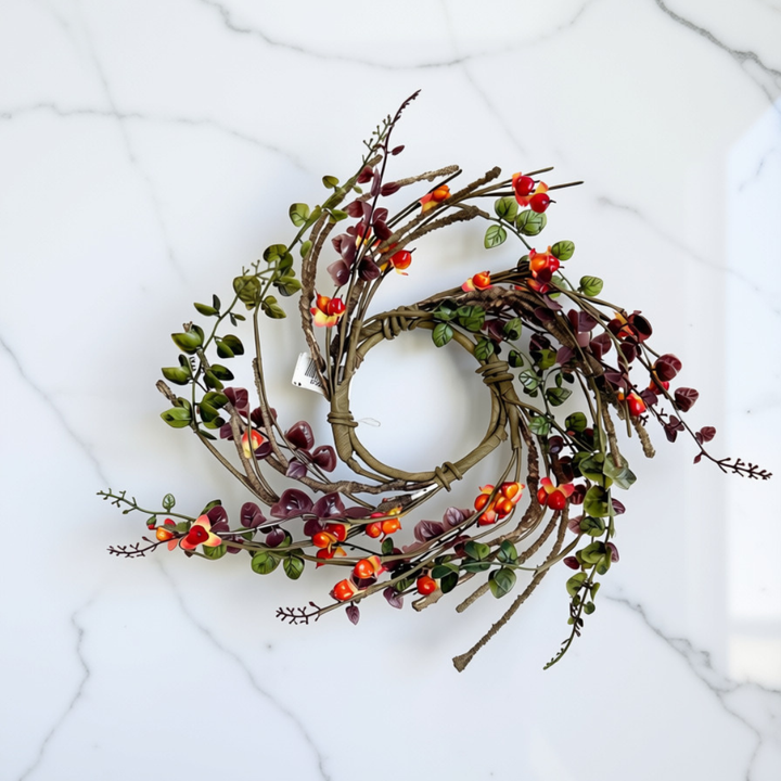 An autumn-themed artificial candle ring with brown twig branches, green and burgundy leaves, and small orange berries, displayed on a white marble surface.