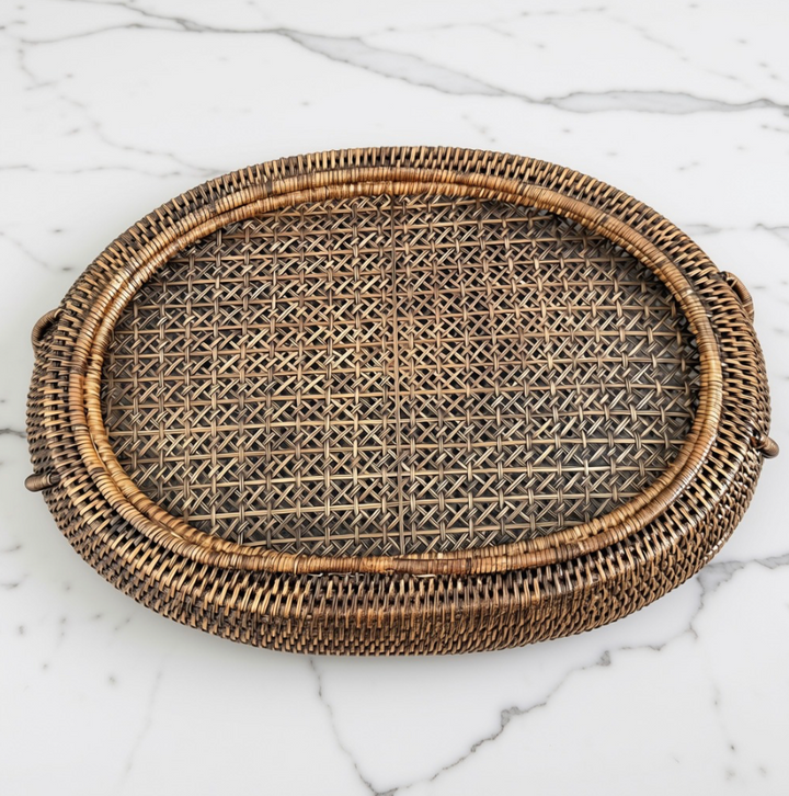 An oval woven rattan tray with a natural brown and beige textured weave, displayed on a white marble surface.
