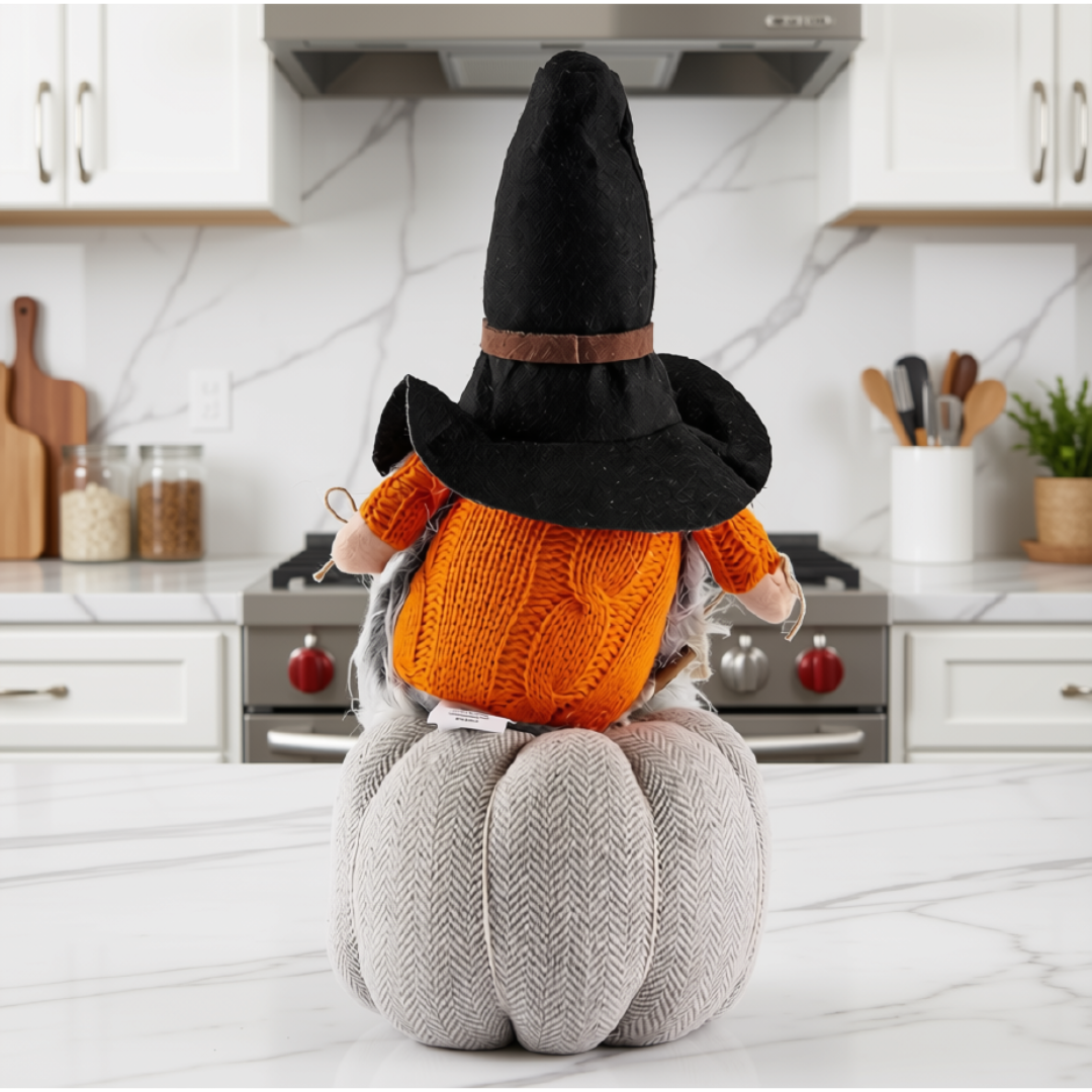 A decorative autumn gnome figurine wearing a black witch hat and orange sweater, sitting on a gray herringbone pumpkin, displayed on a white marble kitchen counter with a stove and cabinets in the background.