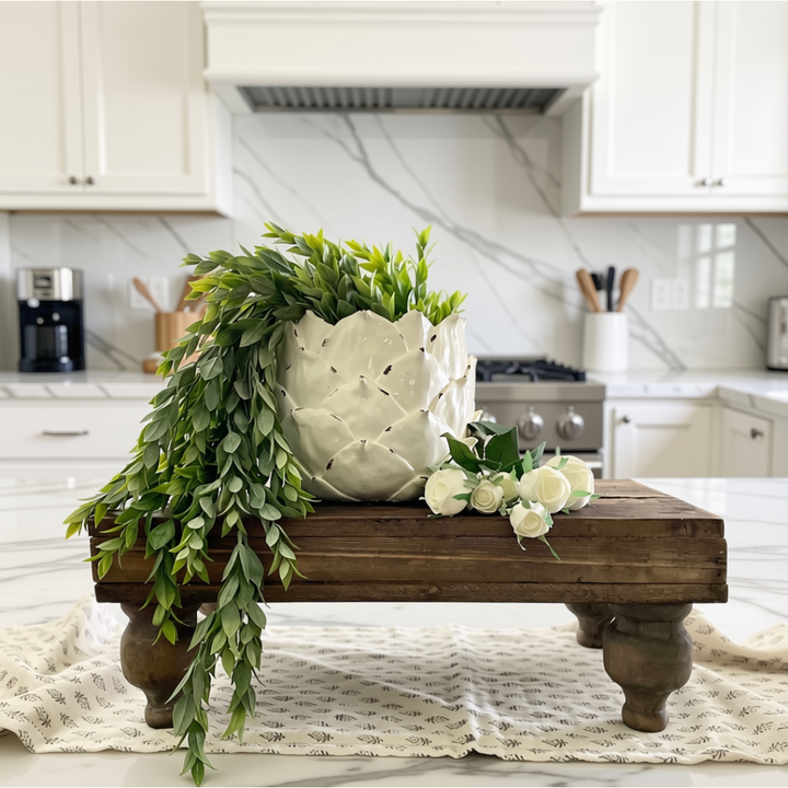 A ceramic planter with a textured, flower-like exterior and a large leafy green trailing plant, displayed on a rustic wooden stand in a bright, modern kitchen setting.