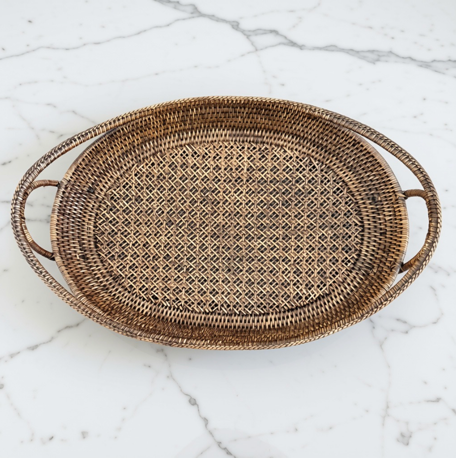 An oval woven rattan tray with two loop handles, shown from a top-down view on a white marble surface.