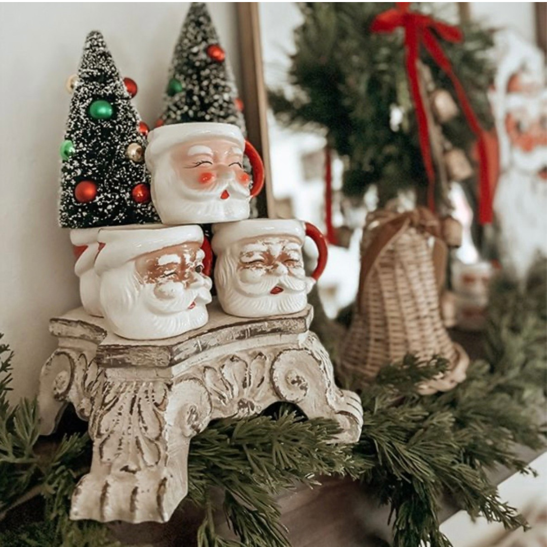 A Christmas-themed decorative set featuring two Santa Claus ceramic heads and two small decorated Christmas trees on an ornate vintage white pedestal riser, styled with green garlands and a bell in the background.