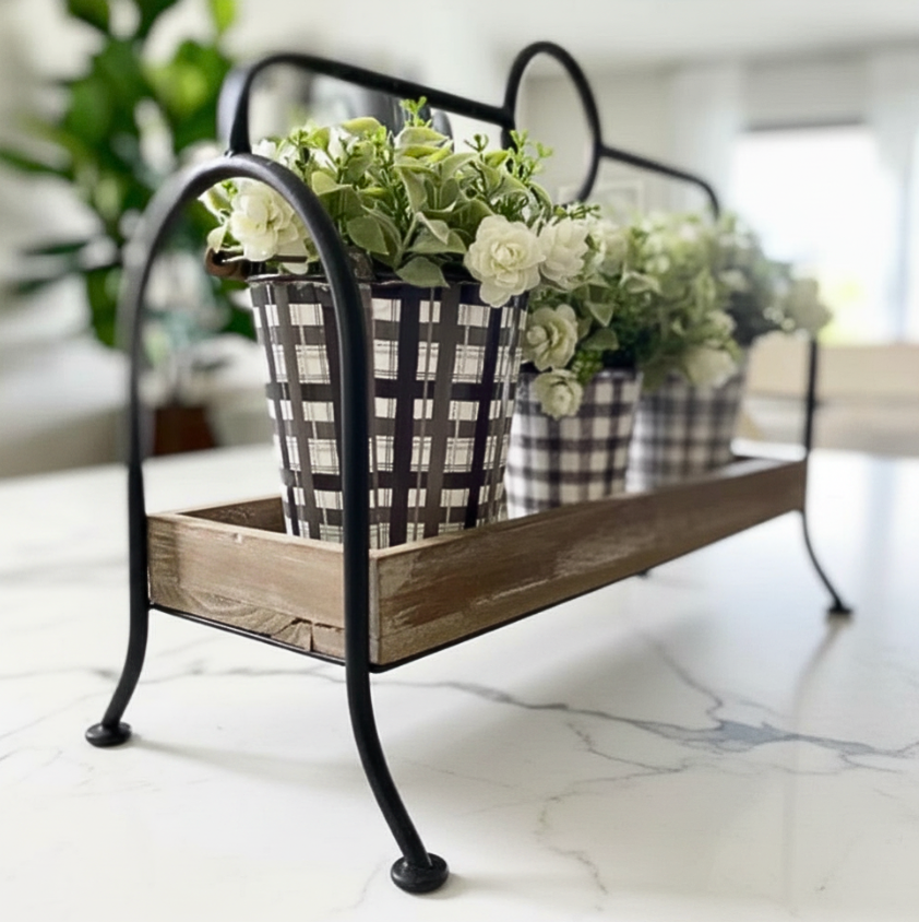 A rustic black metal and wood flower stand holding three black-and-white gingham planters filled with white and green faux flowers, set on a white marble countertop in a bright indoor setting.