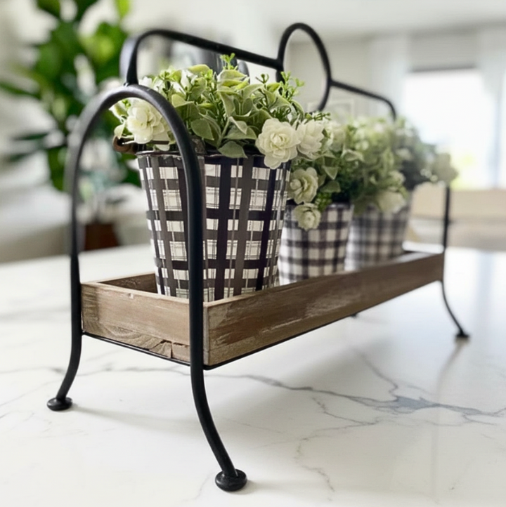A rustic black metal and wood flower stand holding three black-and-white gingham planters filled with white and green faux flowers, set on a white marble countertop in a bright indoor setting.